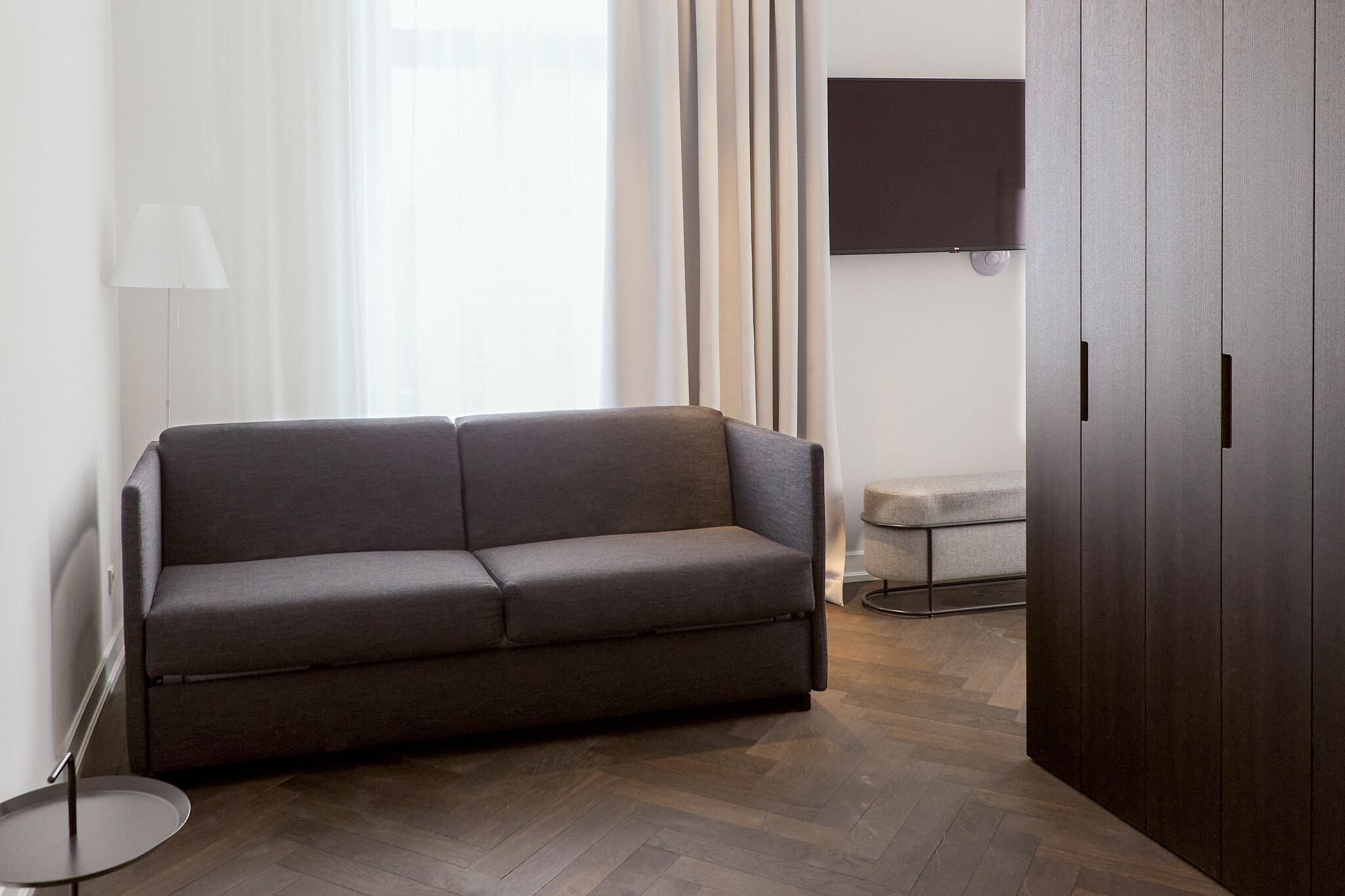 A modern living room with a grey sofa, a round side table, a floor lamp, a TV mounted on the wall, and a dark wooden wardrobe.