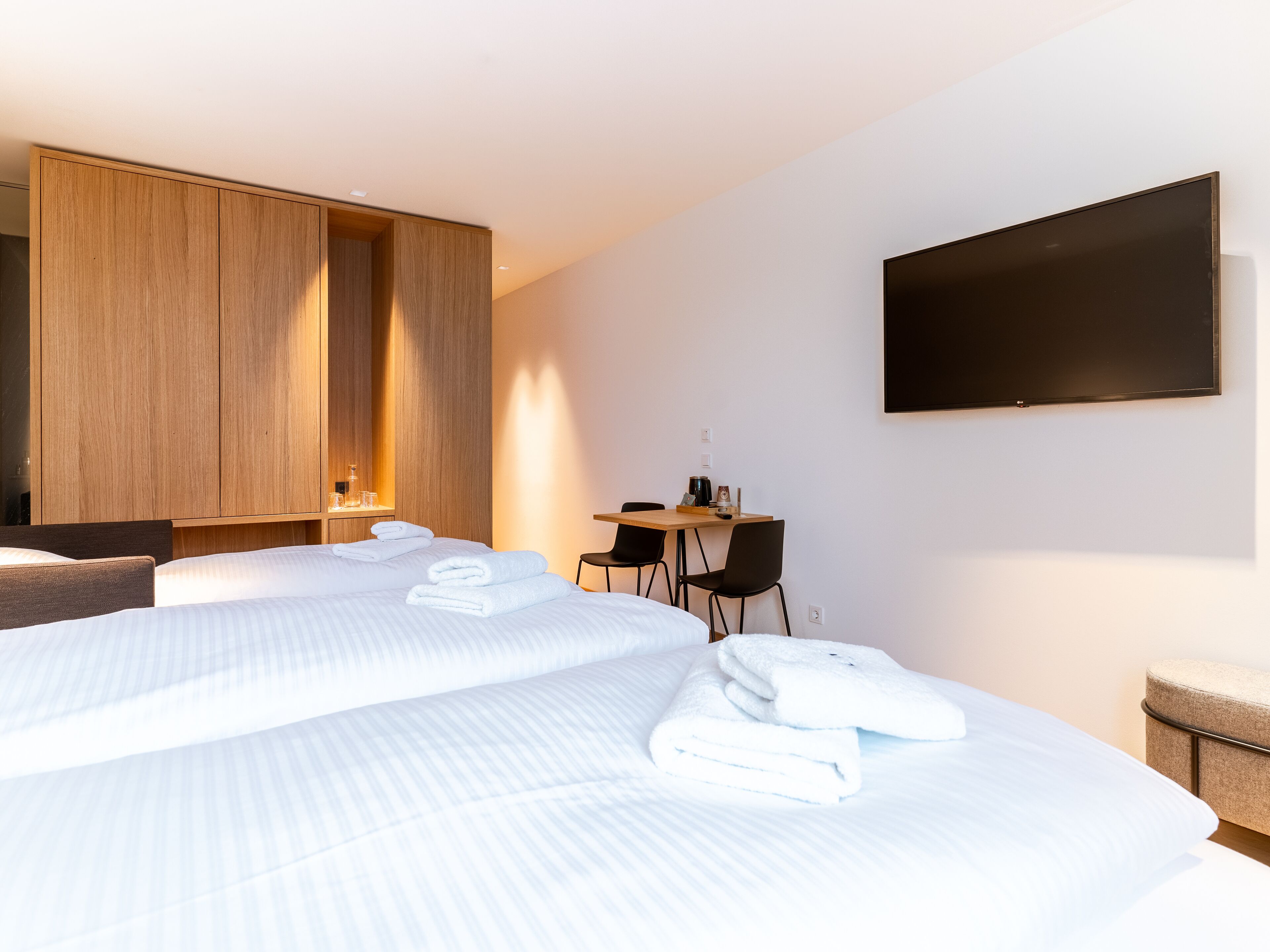 A modern hotel room with two beds, a desk, a chair, a TV, and a wardrobe.