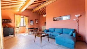Una sala de estar con un sofá azul, vigas de techo de madera, una zona de comedor con sillas y una mesa, y una cocina con encimera y armarios.