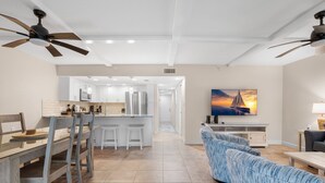 A modern kitchen with a central island, a dining area with wooden chairs, a blue upholstered sofa, and a TV mounted on the wall. - Updated Condo with Intracoastal Views & Direct Vehicle Access – Indian Rocks Beach Retreat (Indian Rocks Beach)