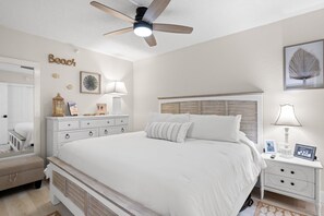 A bedroom with a large bed, a ceiling fan, a nightstand, a dresser, and a wall with decorative items.