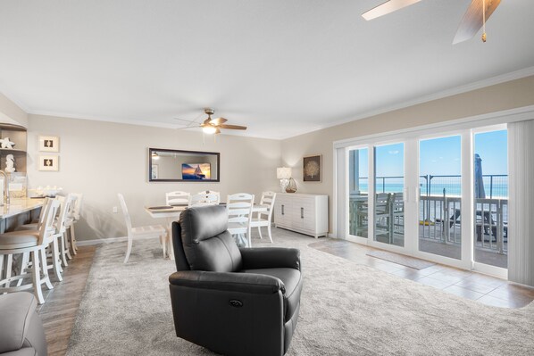 A modern living room with a black leather sofa, a dining area with white chairs, a white cabinet, and a large sliding glass door leading to a balcony with a view.