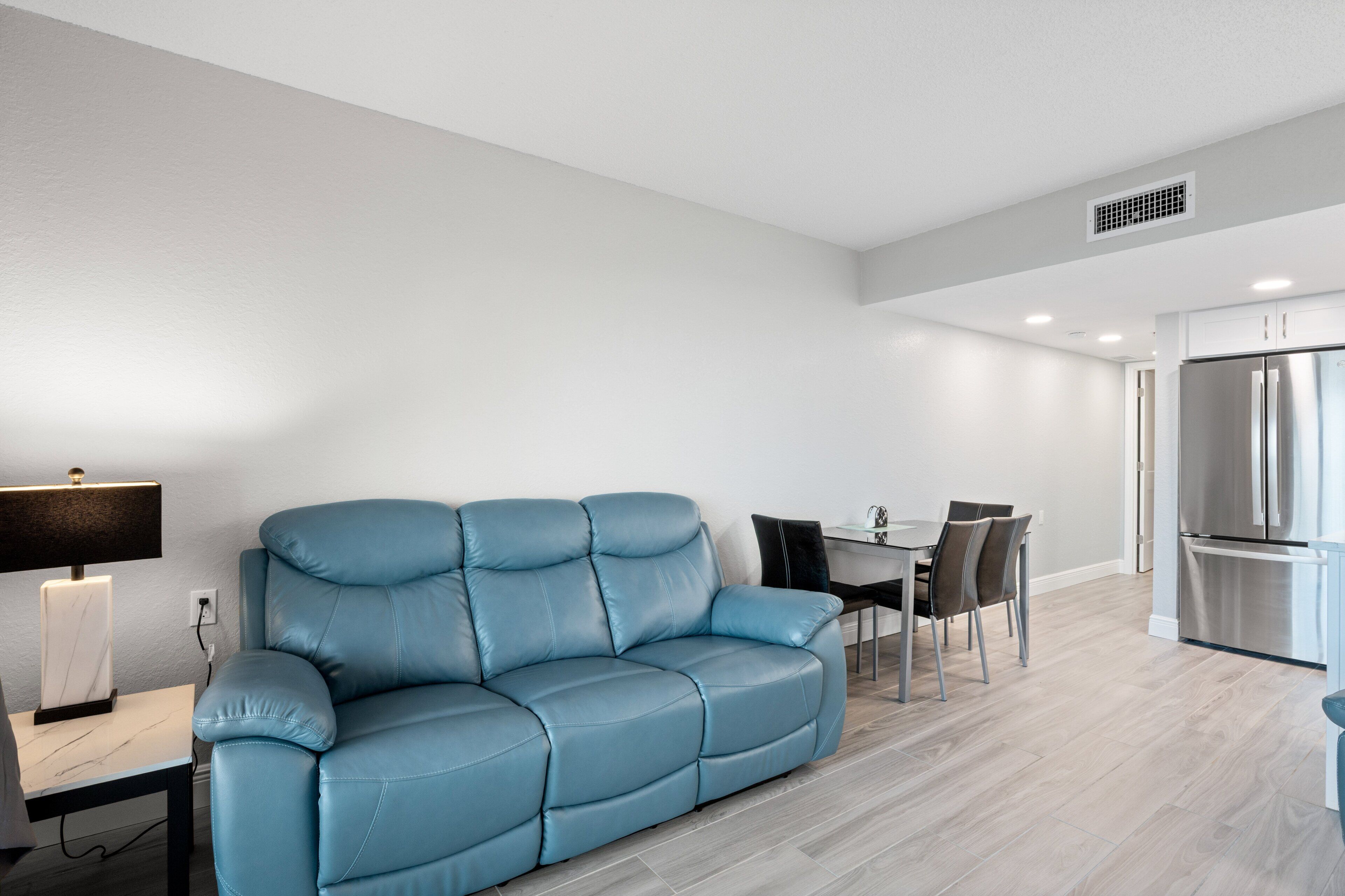Ein modernes Wohnzimmer mit einem blauen Sofa, einem Esstisch mit Stühlen, einer Küchenzeile mit Kühlschrank und einer Lampe auf einem Beistelltisch.