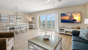 A spacious living room with a large TV, a dining area with white furniture, and a balcony with a view of the ocean. - 🌊 Beachfront 2BR Condo with Direct Pool Access – Steps to the Sand, Sleeps 6 (Indian Rocks Beach)