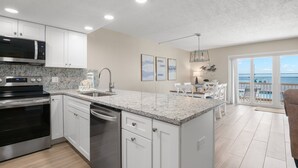 A modern kitchen with white cabinets, granite countertops, stainless steel appliances, and a view of a balcony with outdoor seating. - 🌊 Beachfront 2BR Condo with Direct Pool Access – Steps to the Sand, Sleeps 6 (Indian Rocks Beach)