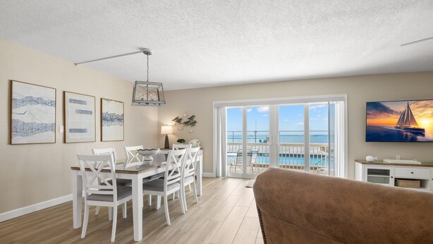 A spacious living room with a large window overlooking a pool, a dining area with white furniture, and a brown couch. - 🌊 Beachfront 2BR Condo with Direct Pool Access – Steps to the Sand, Sleeps 6 (Indian Rocks Beach)