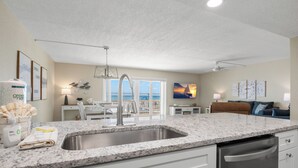 A modern kitchen with a granite countertop, stainless steel sink, and built-in dishwasher. There is a dining area with a white table and chairs, and a view of the ocean through large windows. - 🌊 Beachfront 2BR Condo with Direct Pool Access – Steps to the Sand, Sleeps 6 (Indian Rocks Beach)
