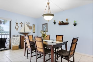 A dining area with a table set for six, surrounded by wooden chairs with woven backs. A chandelier hangs from the ceiling, and wall decorations include a cross, fish, and framed pictures. - π Intracoastal View Condo β Unit #202 2BR/2BA Nautical Charm & Wildlife Views β Sleeps 6 - Indian Rocks Beach (Indian Rocks Beach)