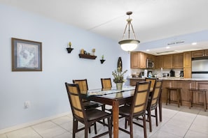 A dining area with a glass-top table surrounded by wooden chairs, a hanging light fixture, and a kitchen in the background. - π Intracoastal View Condo β Unit #202 2BR/2BA Nautical Charm & Wildlife Views β Sleeps 6 - Indian Rocks Beach (Indian Rocks Beach)