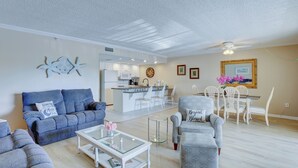 Amplia sala de estar con sofá, sillón y mesa de centro. Se ve una cocina con armarios blancos y un comedor con mesa y sillas.