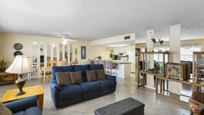 A living room with a blue sofa, a wooden coffee table, and a lamp. There is a kitchen area in the background with a dining table and chairs. - Spacious 2BR Intracoastal View Condo – Discounted Rate  No Balcony, Full Comfort Sleeps 6 (Indian Rocks Beach)