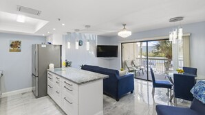 A modern kitchen with a built-in refrigerator, a dining area with a glass table and blue chairs, and a living area with a blue sofa. - Remodeled 1BR Intracoastal View Condo – Designer Touches, Balcony, & Serene Waterway Setting (Indian Rocks Beach)