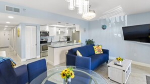 A modern living room with a glass coffee table, blue sofa, and a TV mounted on the wall. - Remodeled 1BR Intracoastal View Condo – Designer Touches, Balcony, & Serene Waterway Setting (Indian Rocks Beach)