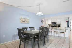 A dining area with a table and chairs, a kitchen in the background, and a clock on the wall.