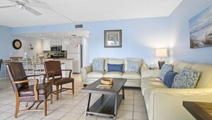A living room with a sofa, two armchairs, a coffee table, a ceiling fan, a kitchen in the background, and a framed picture on the wall. - 🌅 Modern 2BR Intracoastal View Condo – Sunrise Balcony & Serene Comfort on Indian Rocks Beach (Indian Rocks Beach)