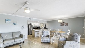 Amplia sala de estar con una mesa de centro de vidrio, un sofá gris y sillones estampados. Se ve una cocina con armarios blancos y una zona de comedor con una mesa y sillas de madera al fondo.