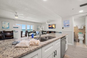 A modern kitchen with a granite countertop, stainless steel sink, and a dishwasher. There are white cabinets, a ceiling fan, and artwork on the walls.