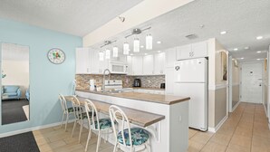 A modern kitchen with white cabinets, a central island with bar stools, a microwave, and a refrigerator. - Coastal Charm & Sunset Views from Your Beachfront Balcony at Fifty Gulfside – Indian Rocks Beach (Indian Rocks Beach)