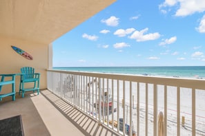 A balcony with a view of the beach, beach chairs, and a surfboard on the wall.