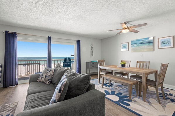 Un salon comprenant un canapé, une table à manger, des chaises et un ventilateur de plafond. On aperçoit, à travers la fenêtre, un balcon donnant sur la plage.