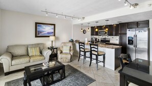 Ein Wohnzimmer mit einer Couch, Sesseln, einem Couchtisch und einer Küchenzeile inklusive Kühlschrank, Mikrowelle und Kochinsel.