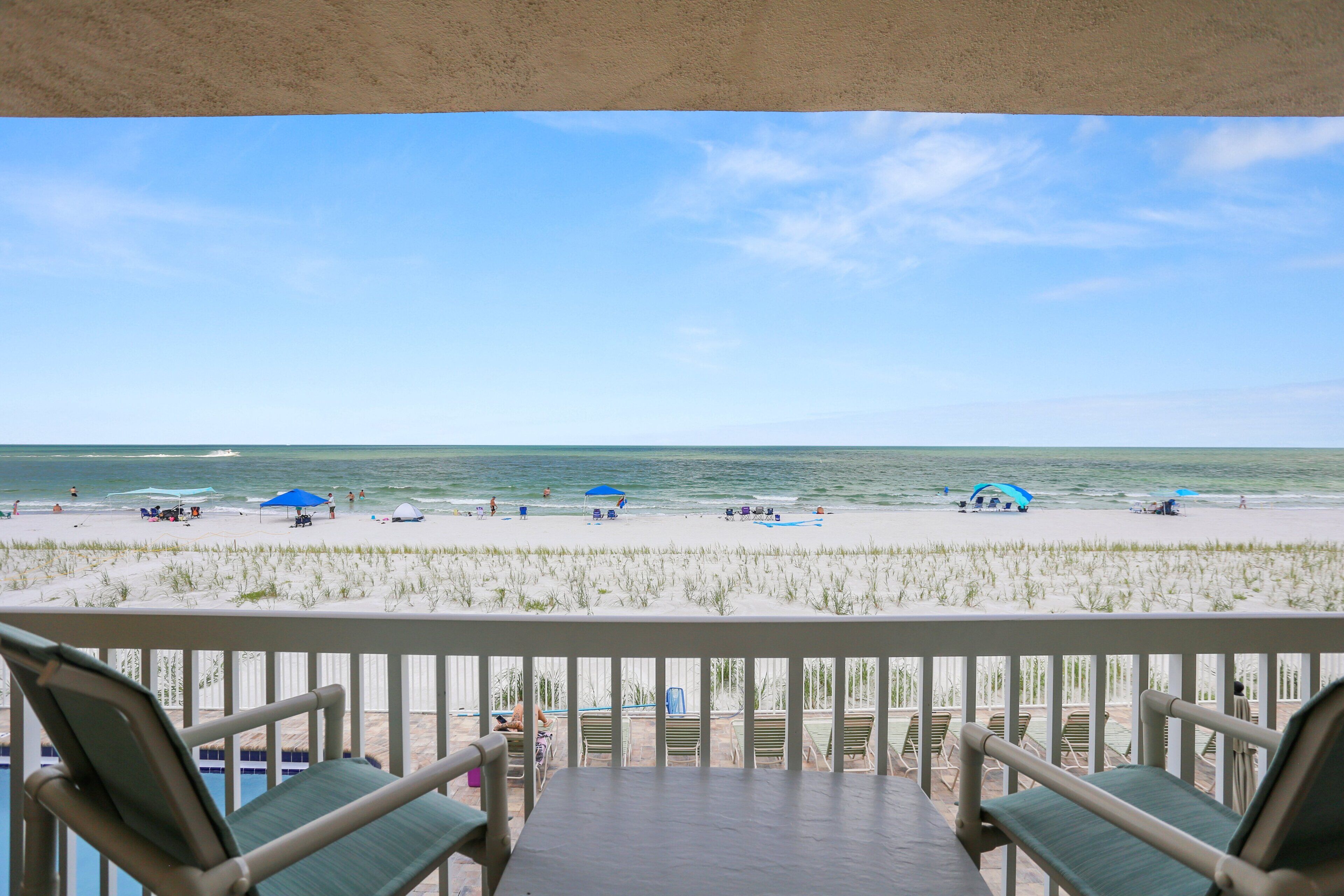 A beach view from a balcony with lounge chairs, a pool, and a clear sky.