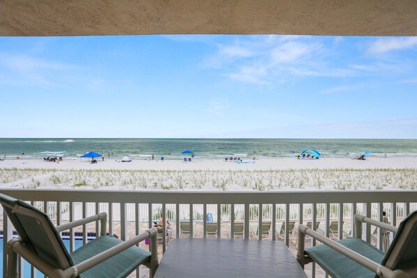 A beach view from a balcony with lounge chairs, a pool, and a clear sky.
