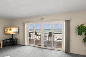 A room with a large sliding glass door leading to a balcony with a view of the ocean. There is a television on the left side of the room, and a plant on the right.