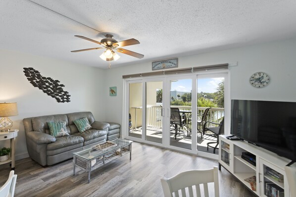A living room with a grey leather sofa, a glass coffee table, a flat-screen TV, and a ceiling fan. - Sunrise Serenity from Your Private Balcony – Renovated Intracoastal Condo 1BR (Indian Rocks Beach)