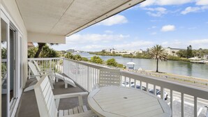 Un balcón con mesa y sillas, con vista a un río y edificios con palmeras al fondo.