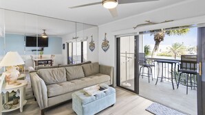 Una sala moderna con sofá, otoman y ventilador de techo. Hay un comedor con mesa y sillas, y un balcón con mobiliario exterior.