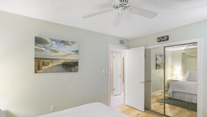 Un dormitorio con una cama, un cuadro de una escena de playa, un ventilador de techo y una puerta corredera que da al baño.