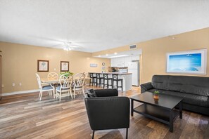 A living room with a black sofa, a dining area with white chairs, a kitchen with white cabinets, and a ceiling fan.