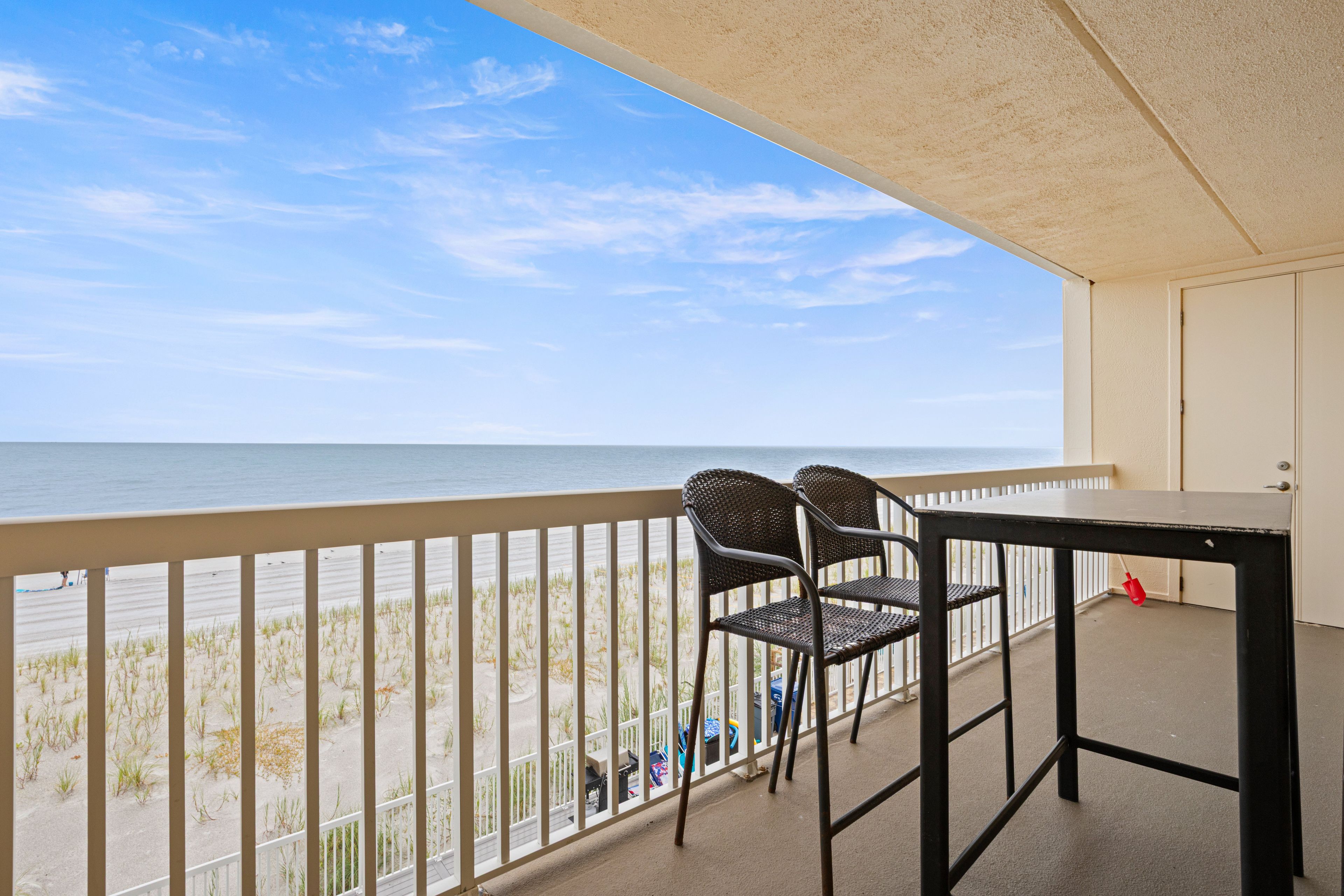 A balcony with a view of the beach, two wicker chairs, and a table.
