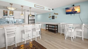 Une cuisine moderne avec des meubles blancs, un dosseret en marbre et des luminaires suspendus. On y trouve un coin repas avec des chaises blanches et une table en bois. Un téléviseur fixé au mur diffuse une scène de coucher de soleil.