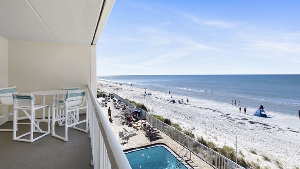 Ein Balkon mit Pool, weißen Möbeln und Blick auf einen Strand mit Personen und einem Zelt.