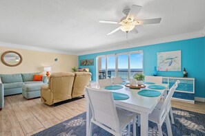 A living room with a sofa, a dining table with chairs, a ceiling fan, and a view of the ocean.