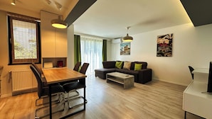 A modern living room with a wooden dining table, a sofa, and a coffee table. There are two pendant lights, a radiator, and a window with blinds. - Timisoara Prime Residence | powered by NEO (Timisoara)