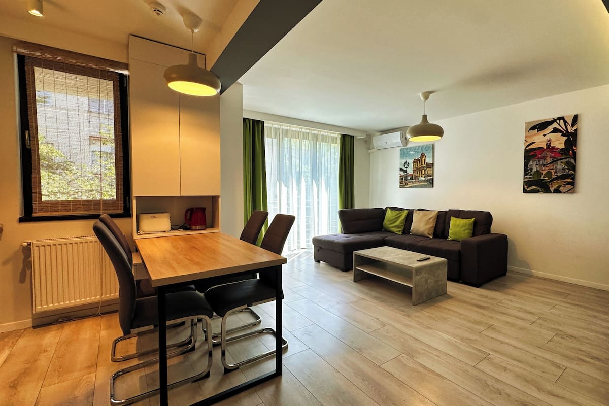 A modern living room with a wooden dining table, a sofa, and a coffee table. There are two pendant lights, a radiator, and a window with blinds.