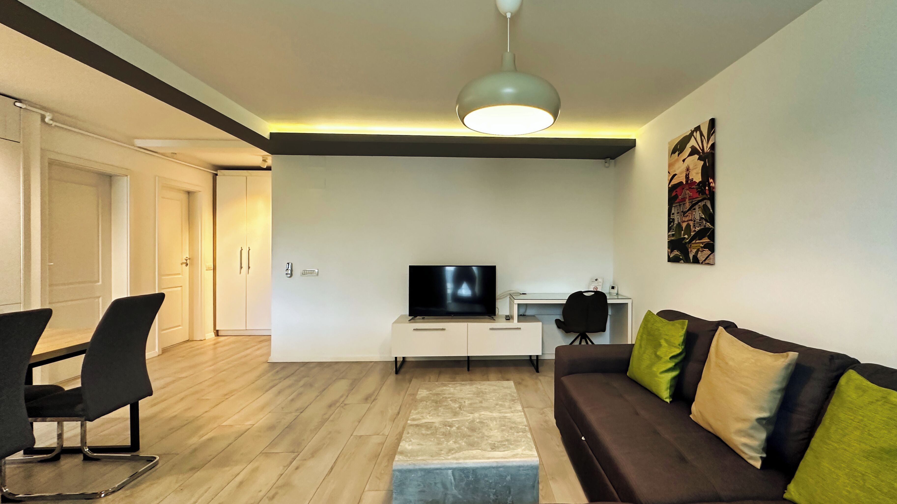 A modern living room with a flat-screen TV, a sofa with cushions, a dining area with chairs, and a desk with a chair.