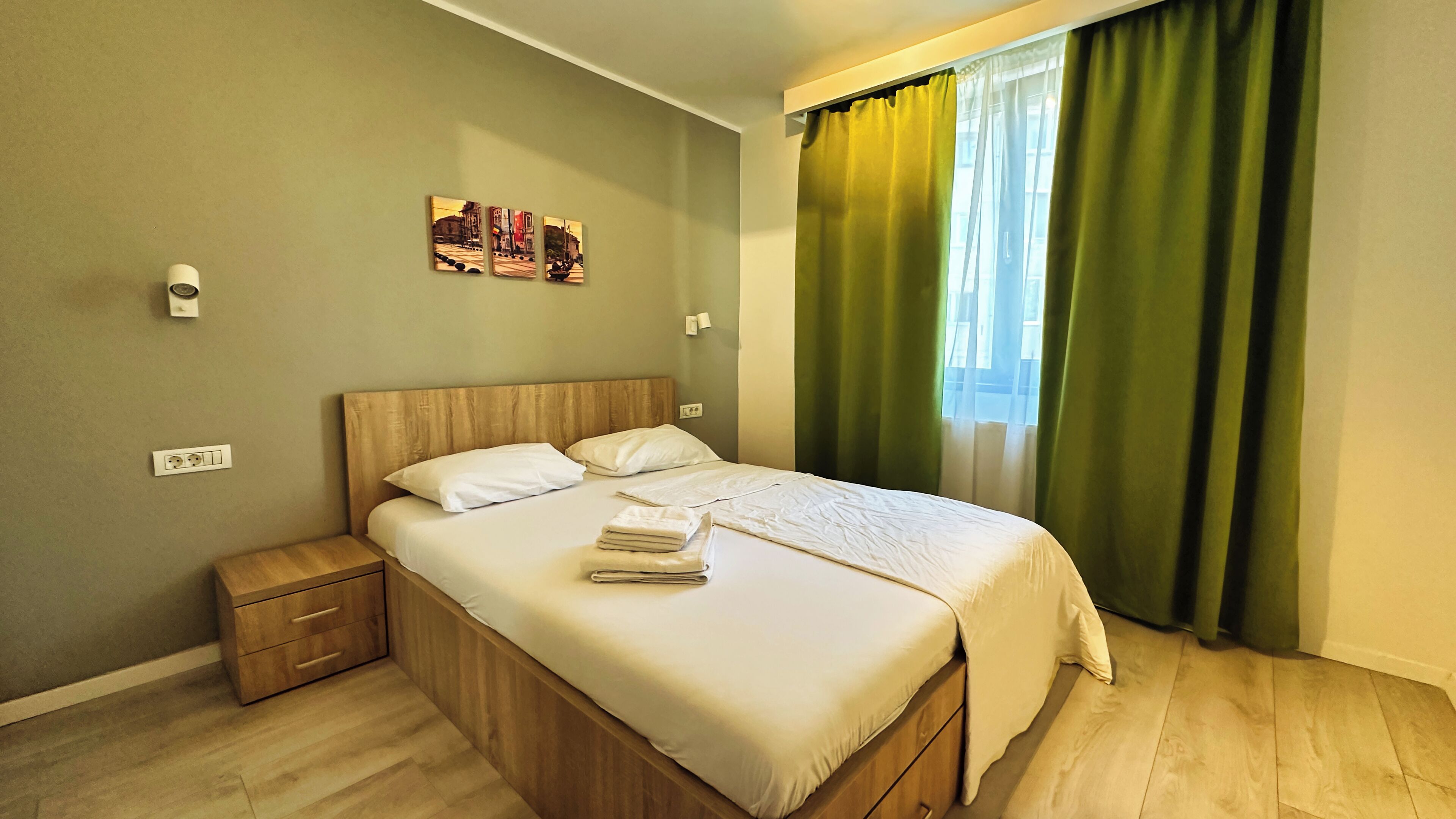 A neatly made bed with a wooden headboard, a bedside table with a drawer, and a power outlet on the wall.