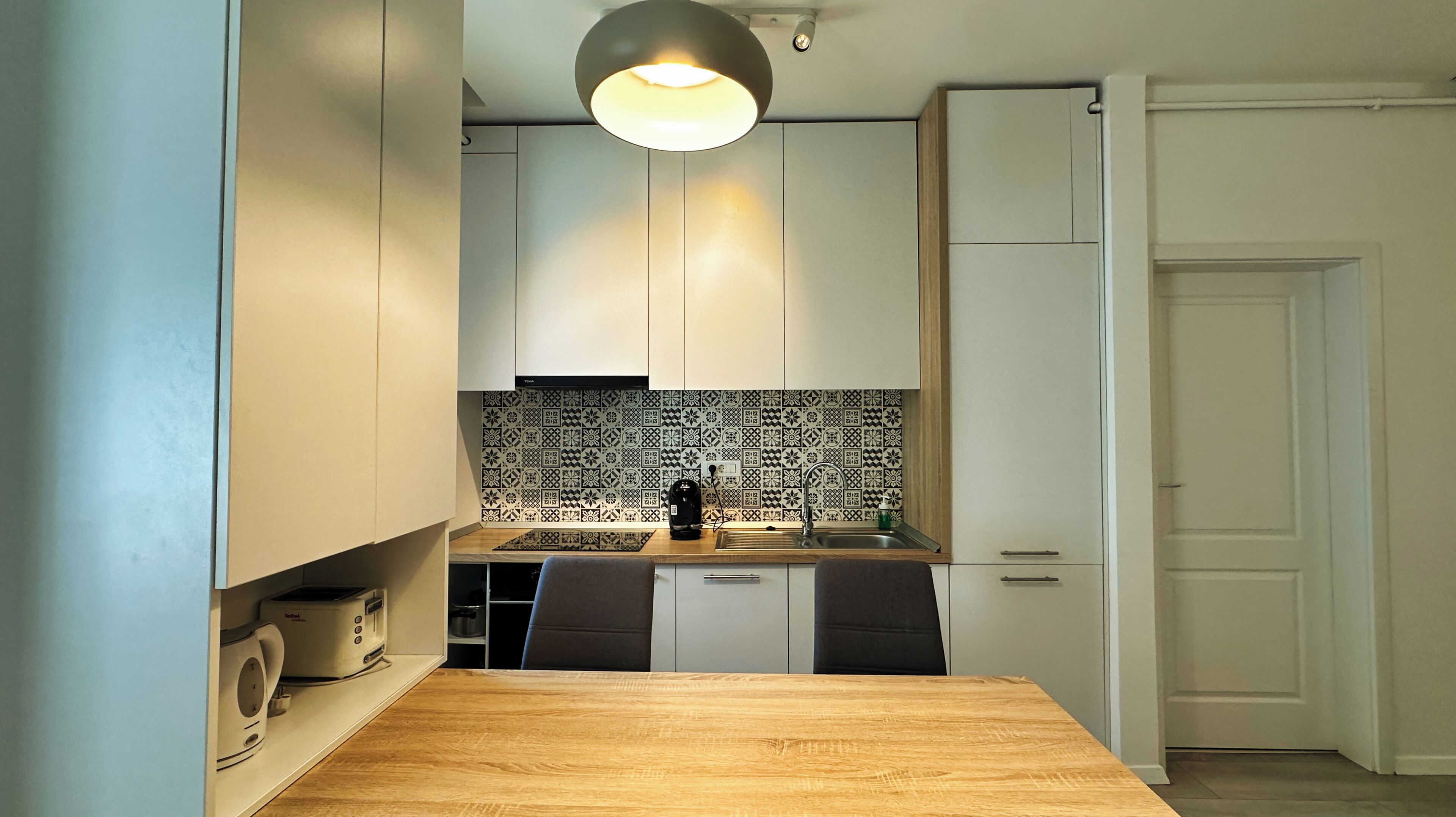 A compact kitchen with white cabinets, a wooden countertop, a patterned backsplash, and a single pendant light.