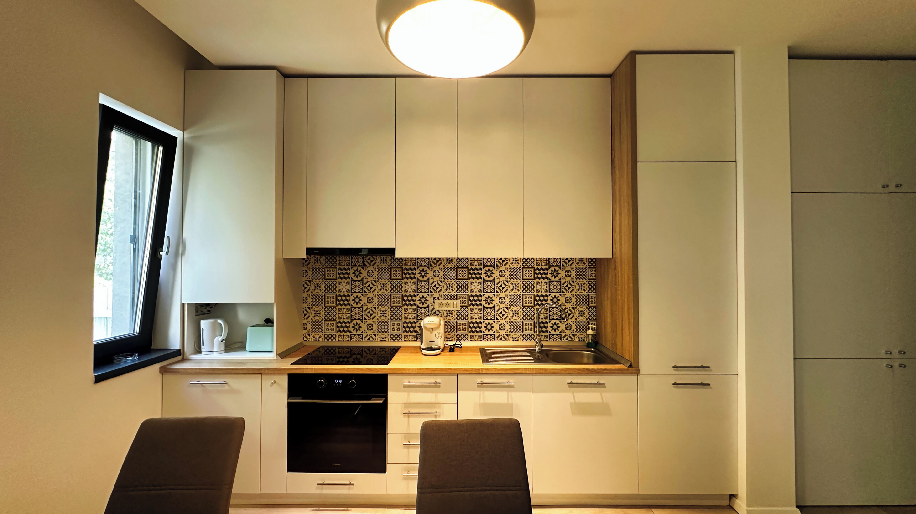 Cocina compacta con estufa negra, cafetera y pileta. Una ventana a la izquierda y un mueble a la derecha.