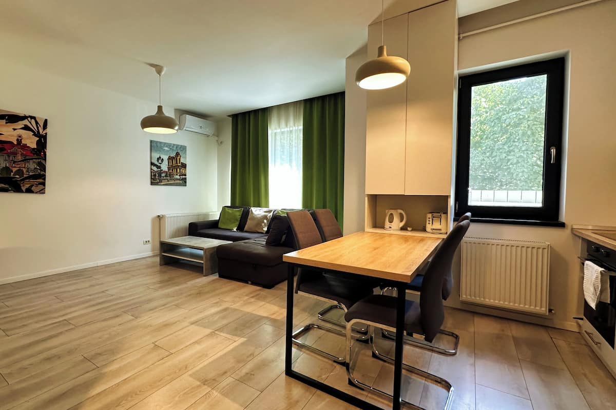 A modern living room with a wooden floor, a sofa, a dining table, and a kitchen area with a window.