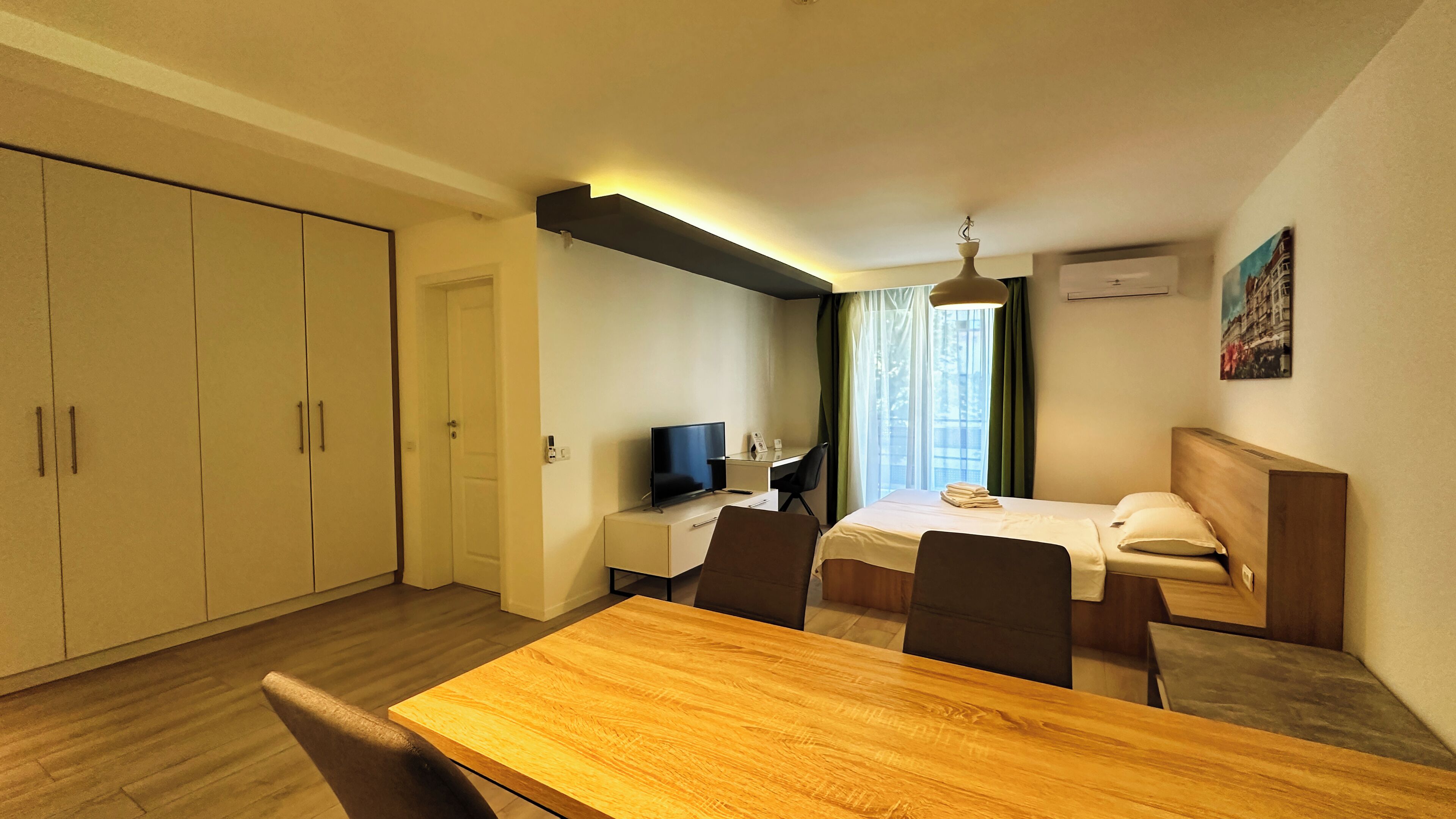 Ein Hotelzimmer mit einem Holztisch, einem Bett mit weißer Bettwäsche, einem Schreibtisch mit Fernseher und einem Kleiderschrank.