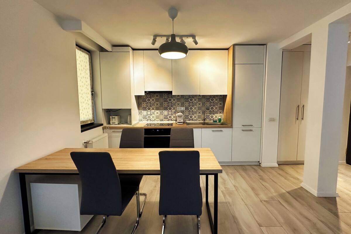 A modern kitchen with a dining area, featuring a wooden table, two chairs, a built-in oven, and a patterned backsplash.
