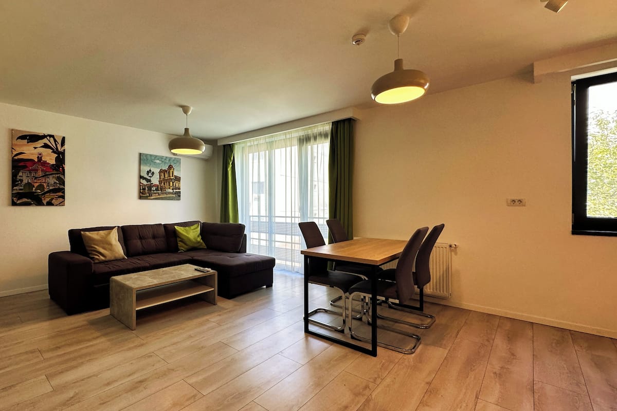 A modern living room with a sectional sofa, a dining table with chairs, and a large window with curtains.