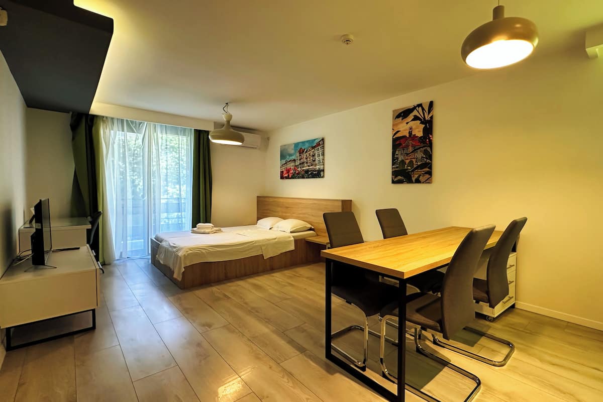 A modern hotel room with a wooden bed, a desk with a computer, a dining table with chairs, and a large window with curtains.