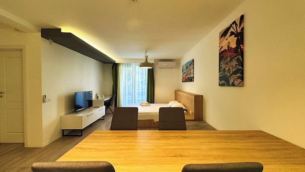 A modern hotel room with a wooden dining table, a bed with a wooden headboard, a desk with a computer and a TV, and two chairs. - Timisoara Prime Residence | powered by NEO (Timisoara)
