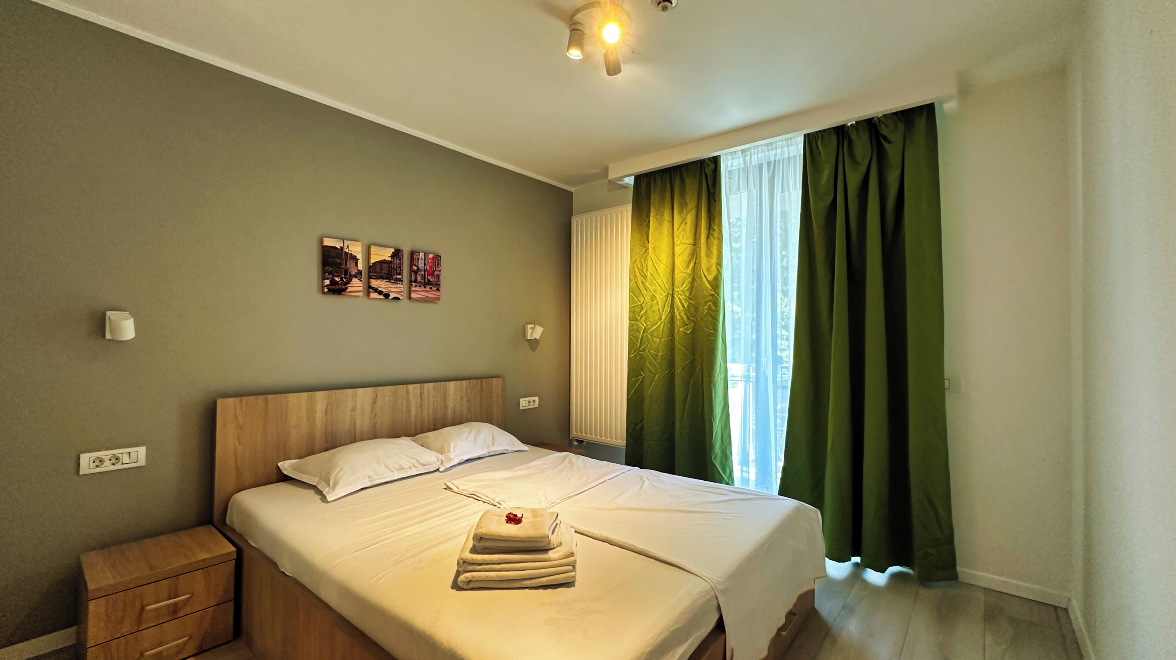 A neatly made bed with a white comforter and pillows, a wooden nightstand, a wall-mounted light, and a window with green curtains.